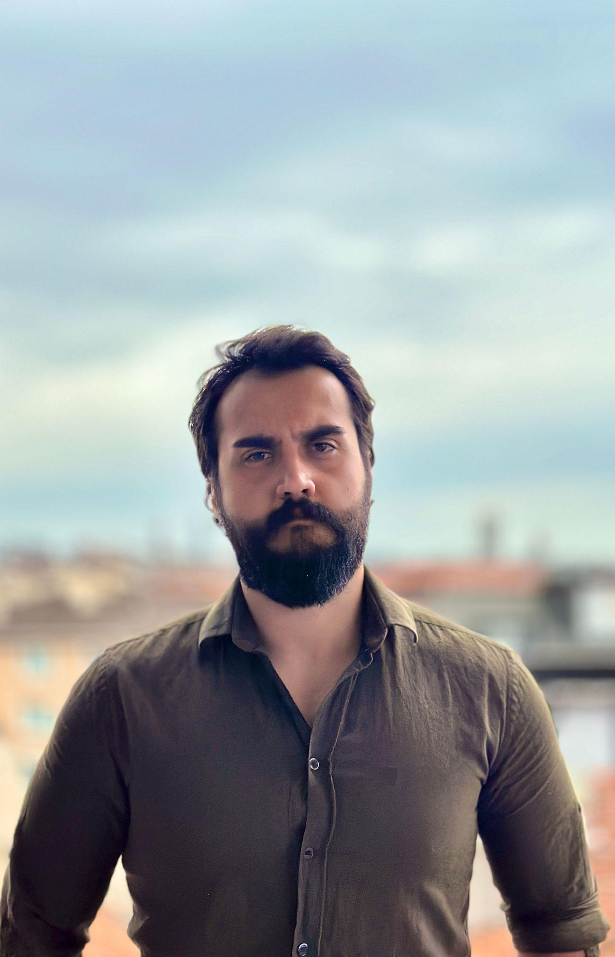 Person in a button-up shirt standing outdoors with a cloudy sky in the background.
